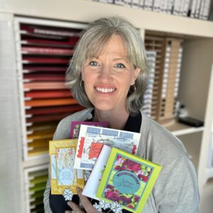 Sarah Storebo, independant Stampin' Up! Demonstrator and card crafter, holding a variety of cards in front of a shelf of cardstock, ink, and stamps